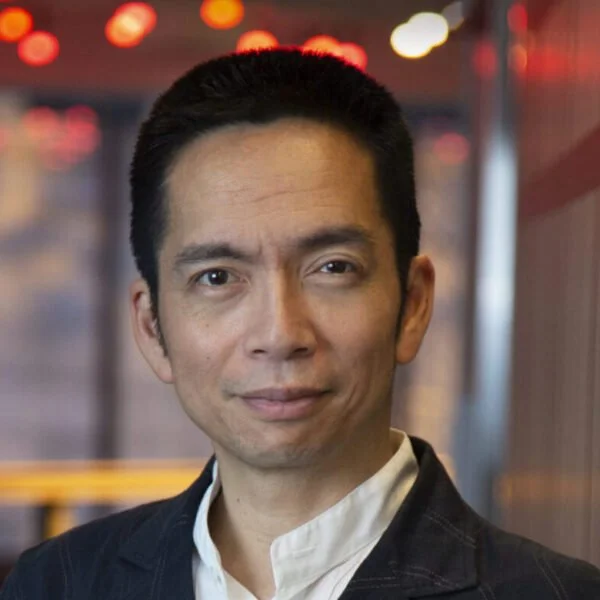 A man with short black hair, wearing a white shirt and dark jacket, smiling in an indoor setting with colorful bokeh lights in the background.
