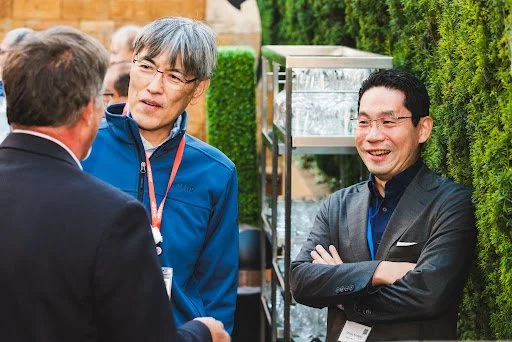 Four people are engaged in conversation outdoors, with two men in the foreground talking to each other, one wearing a blue jacket and glasses, the other a black suit. Another man, also in glasses and a black jacket, stands nearby smiling and listening.