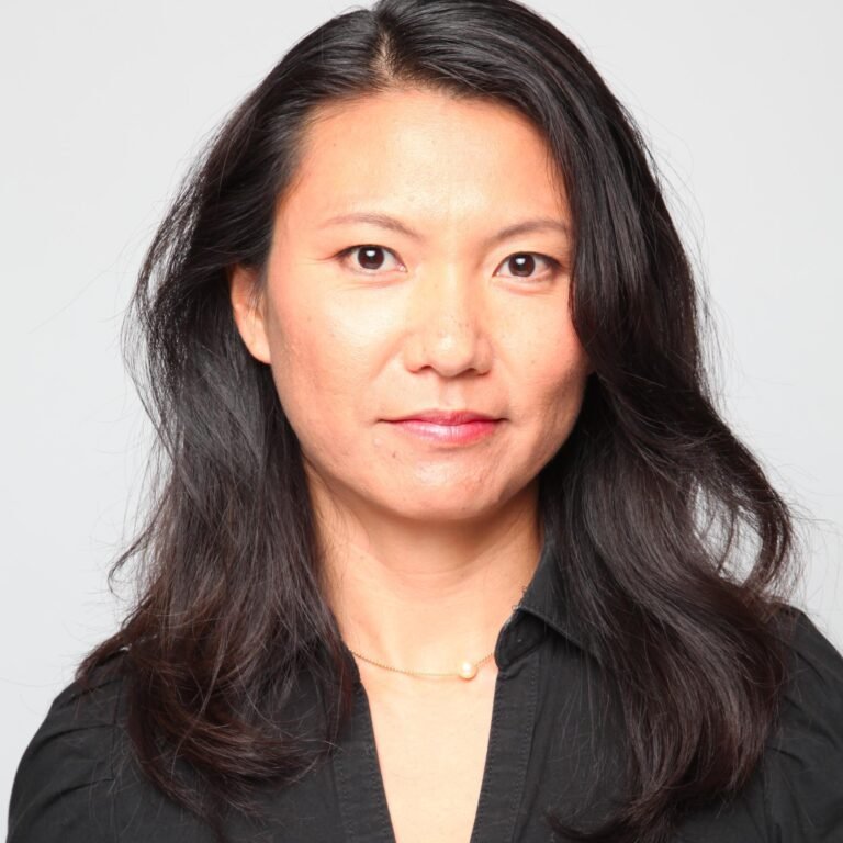 A woman with shoulder-length black hair wearing a black top and a pearl necklace, posing against a plain light grey background.