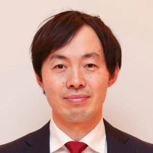 A man with dark brown hair wearing a suit and tie, standing against a plain light-colored background.