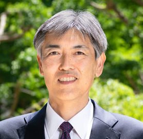 A man with gray hair wearing a suit and tie smiling outdoors surrounded by greenery.
