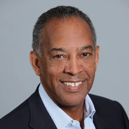 Professional headshot of a middle-aged African American man with short gray hair, smiling in a dark suit jacket and light blue shirt against a gray background.