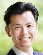A smiling man with black hair wearing a suit, outdoors with a blurred green background.
