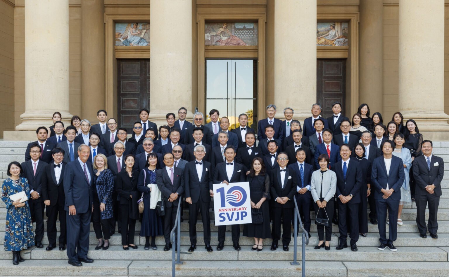 Group of formally dressed men and women standing on the steps of a building, holding a sign that reads '10th Anniversary SVJP'.