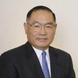A middle-aged man wearing glasses, a dark suit, white shirt, and blue tie, standing against a plain background.