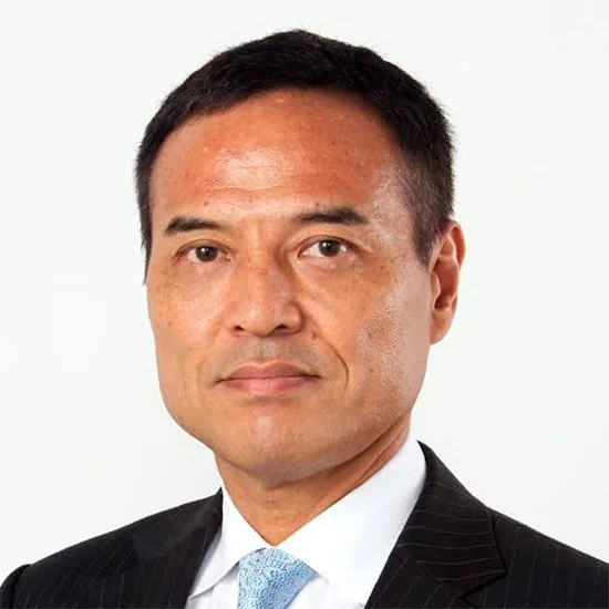 Portrait of a man in a black suit and light blue tie, with short black hair and a serious expression, against a plain white background.