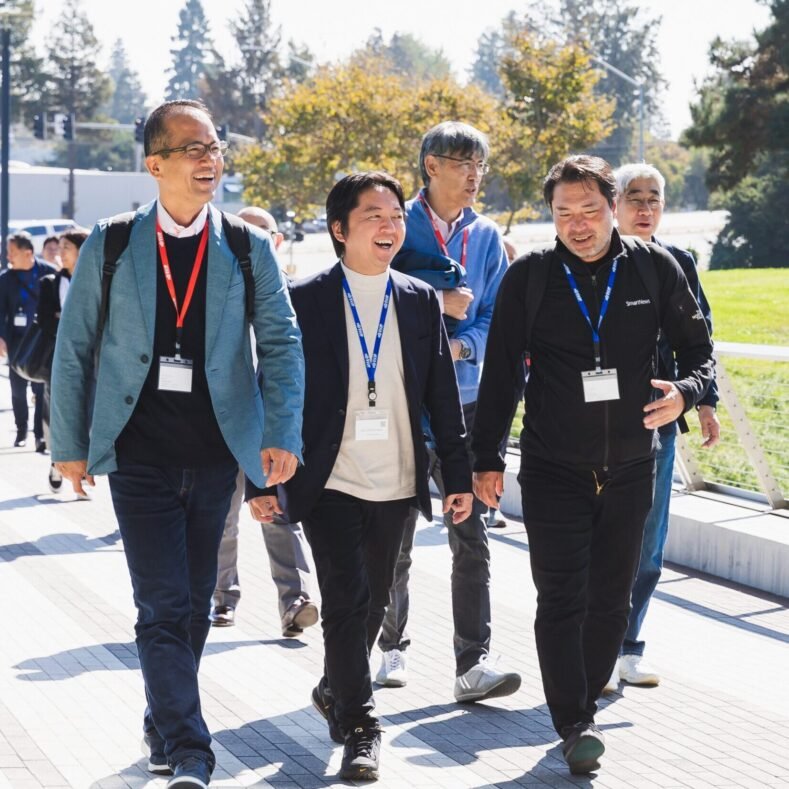 Group of five smiling men walking outdoors on a pathway, wearing conference badges and casual to business attire, with trees and a grassy area in the background.