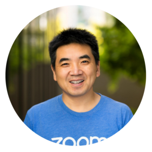 A man smiling outdoors, wearing a blue shirt with 'Zoom