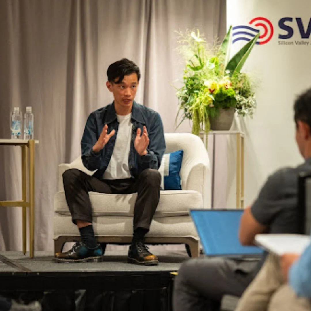 Man sitting on a cream-colored sofa speaking during a panel discussion or interview at Silicon Valley Summit, with a person using a laptop in the foreground and a floral arrangement behind him.