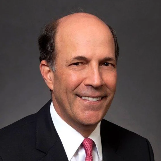 Close-up portrait of smiling middle-aged man with light skin, wearing a dark suit, white shirt, and pink tie, against a gray background.