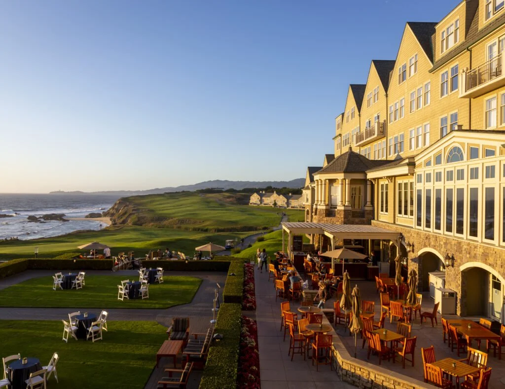 Outdoor patio dining area at sunset with orange chairs and umbrellas, overlooking a golf course and ocean cliffs, adjacent to a large yellow building with multiple stories and numerous windows.
