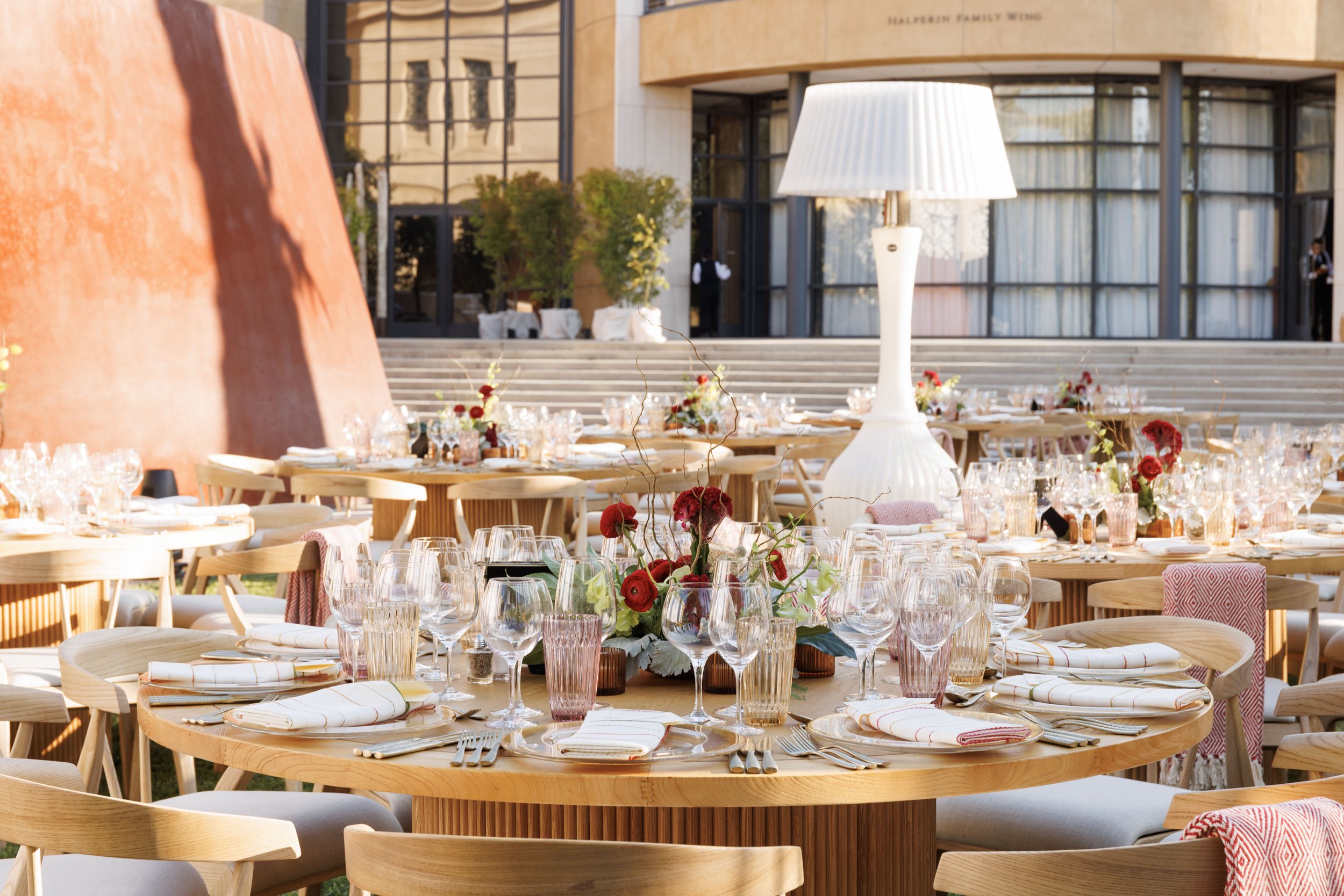 Outdoor event setup with round wooden tables decorated with red flowers and glassware, set in front of a modern building with large glass windows and a large white floor lamp as a centerpiece.