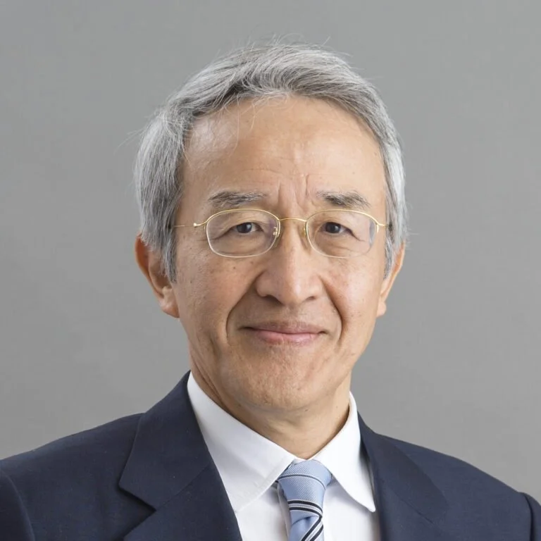A middle-aged man with gray hair, glasses, wearing a dark suit, white shirt, and a striped tie, smiling softly against a plain gray background.