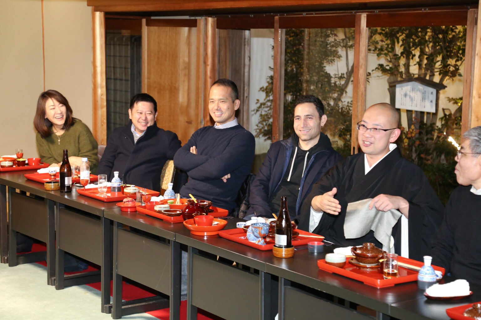 Group of six people sitting at a long dining table, smiling and engaging in conversation, in a restaurant with wooden decor and large windows showing an outdoor garden.