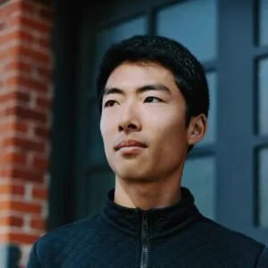 A young man with short black hair wearing a dark zip-up jacket, standing outdoors in front of a building with brick and window features.