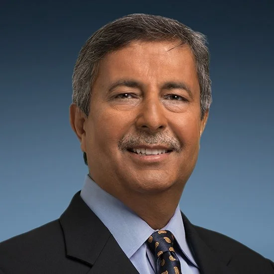 Headshot of a middle-aged man with gray hair, a mustache, wearing a dark suit, light blue shirt, and a patterned tie, smiling against a gray gradient background.