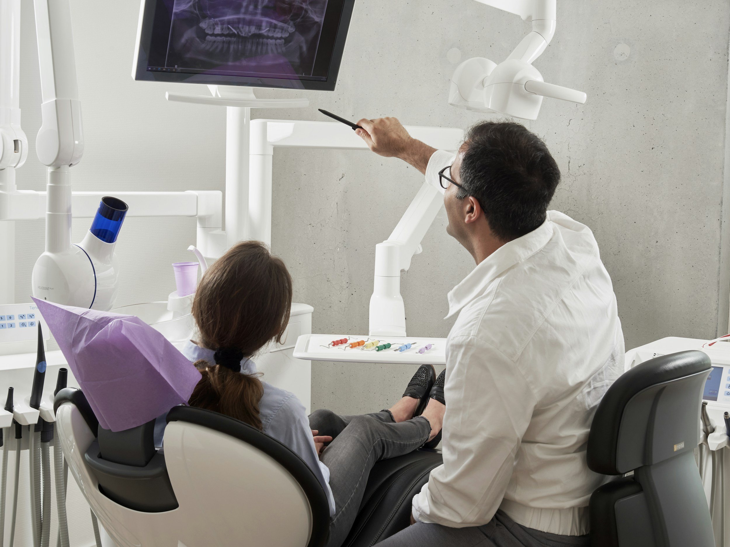 A dentist examining a young girl's teeth in a dental clinic, with dental equipment and a monitor displaying an x-ray in the background.
