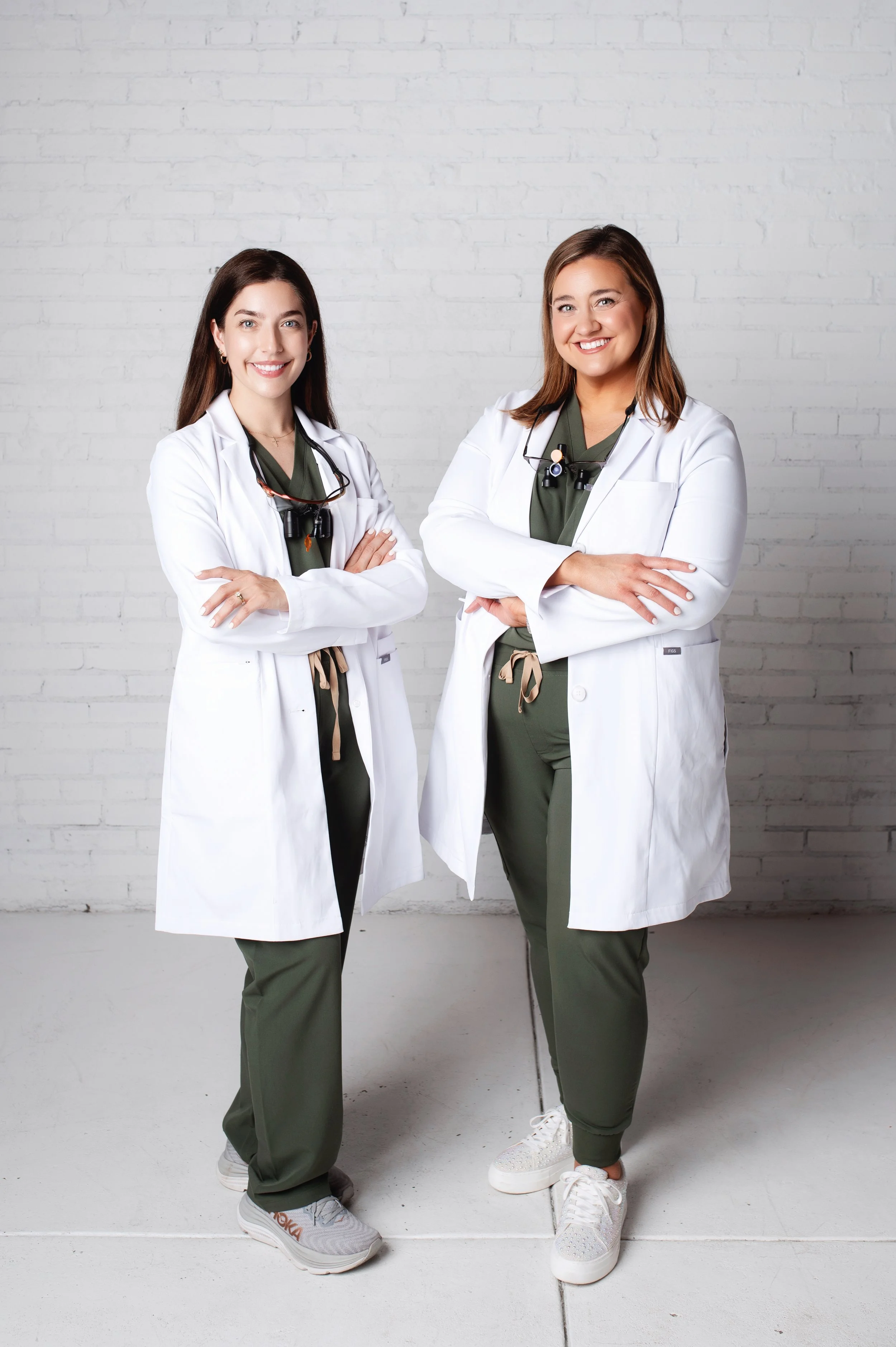 Two female healthcare professionals standing side by side in scrubs and white lab coats, smiling, with arms crossed against a white brick wall background.
