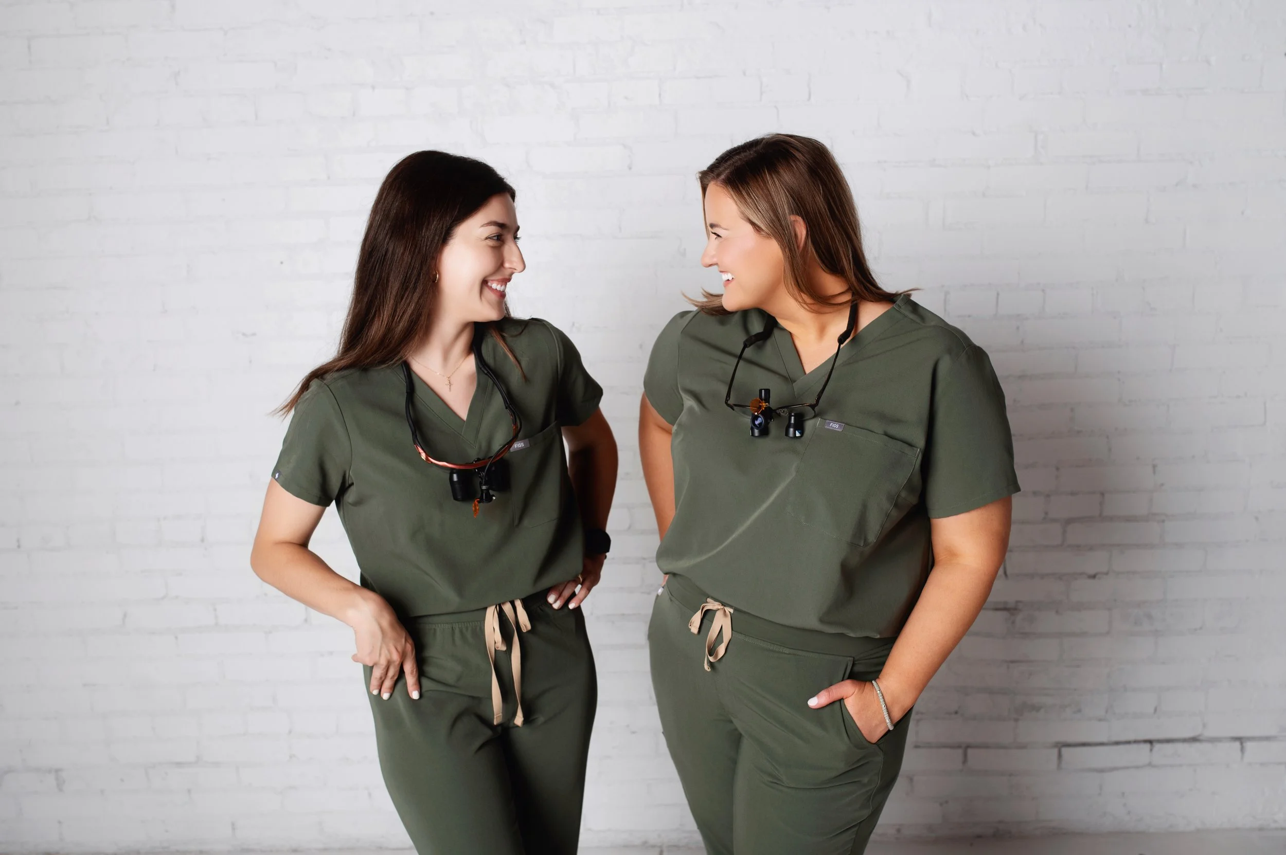 Two women in green scrubs facing each other and smiling, standing against a white brick wall. They are wearing stethoscopes and medical badges around their necks.
