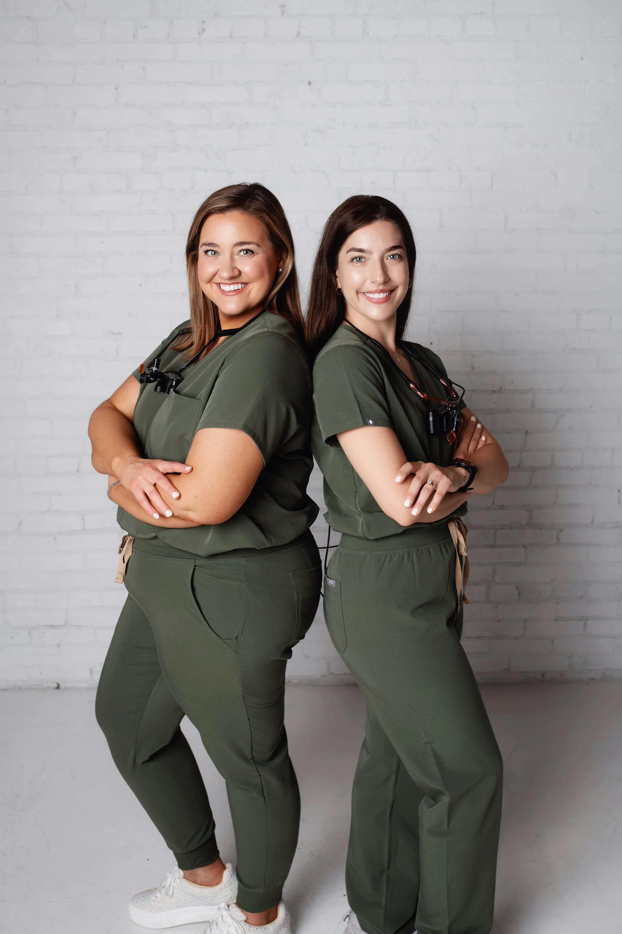 Two women dressed as healthcare workers stand back-to-back with crossed arms in front of a white brick wall, smiling.