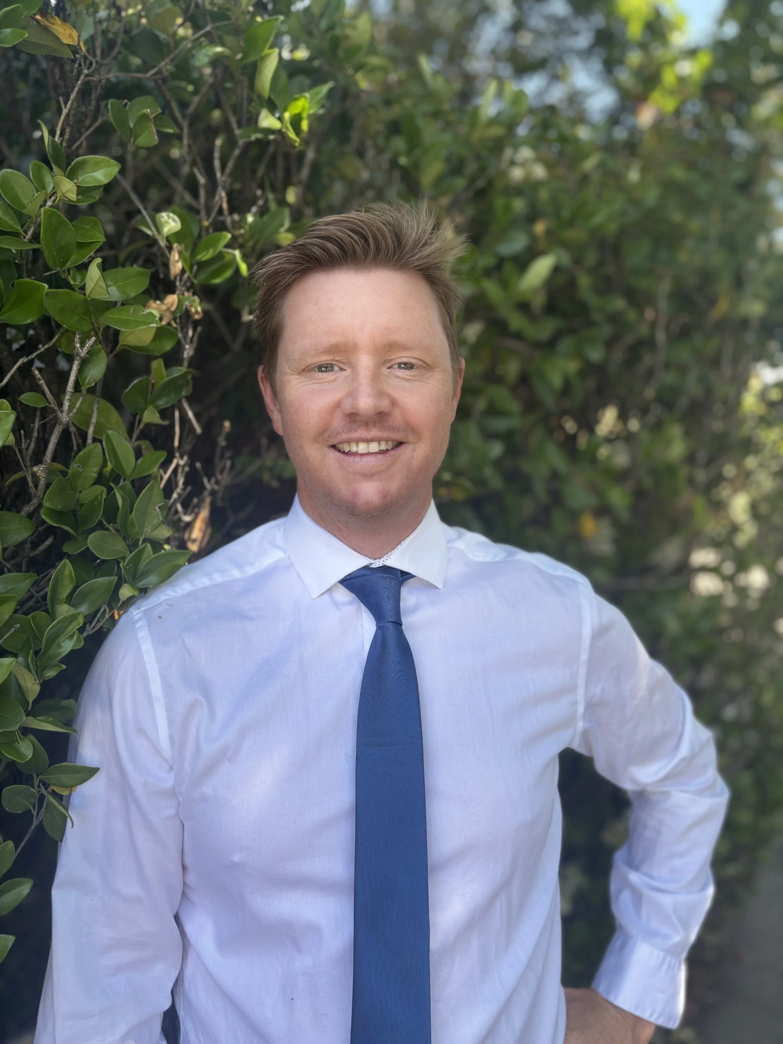 A man in a white shirt and blue tie smiling outdoors in front of green foliage.