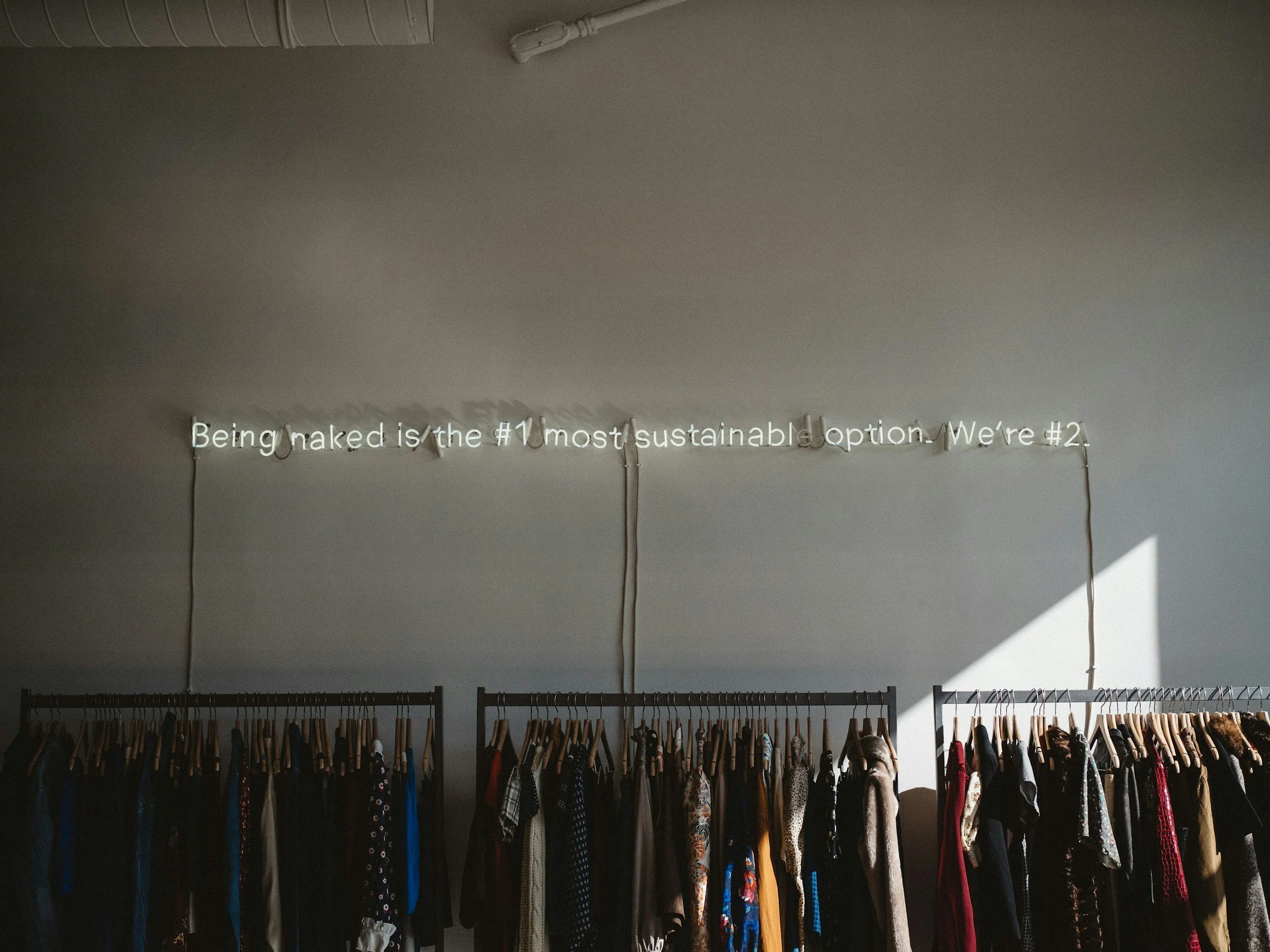 Clothing racks with various dresses and garments in a clothing store or boutique, with a neon sign in the background that reads 'Being naked is the #1 most sustainable option. We're #2.' in white lights.