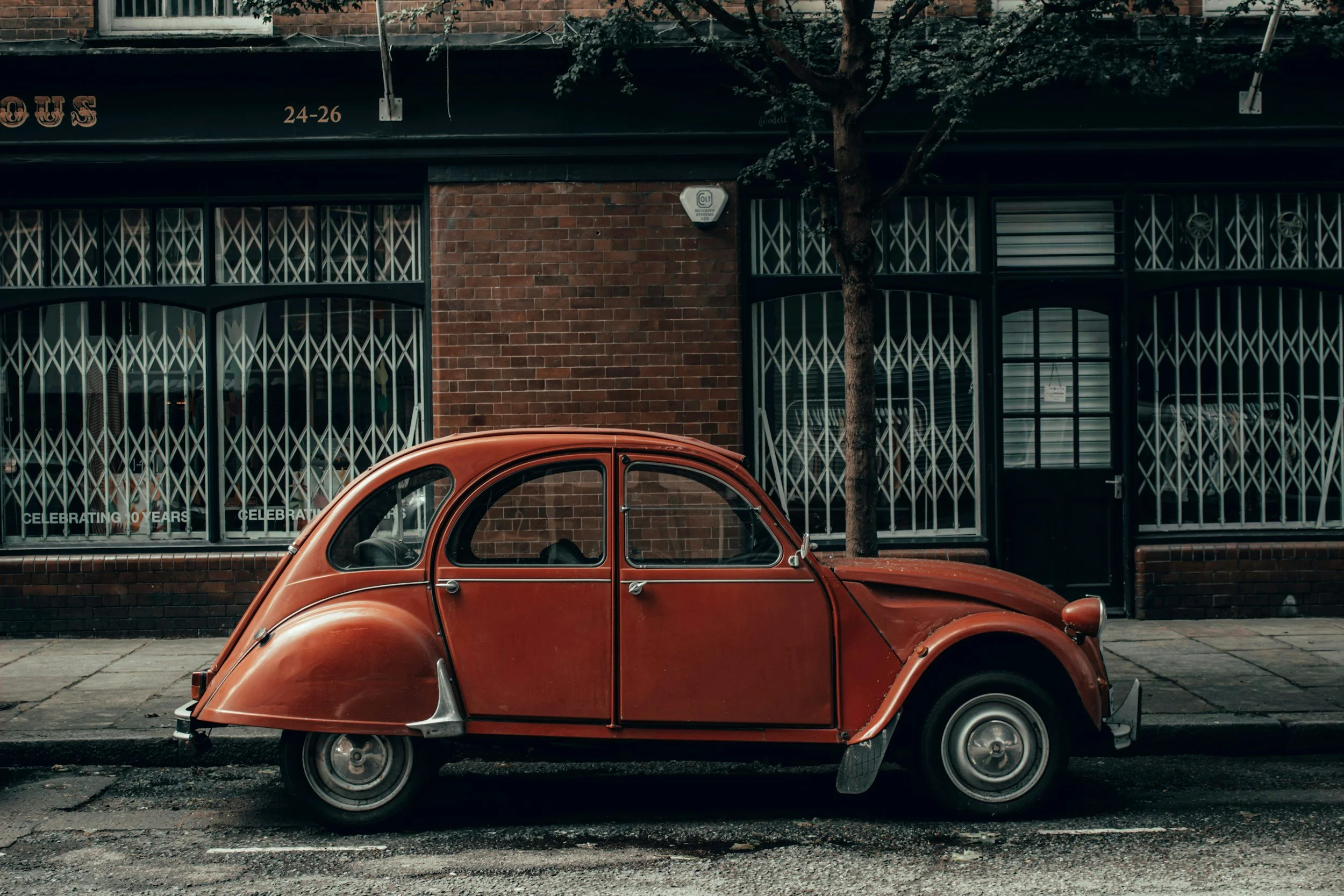 A vintage orange car parked on a street in front of a brick building with barred windows and a sign, with a tree partially obscuring the building.