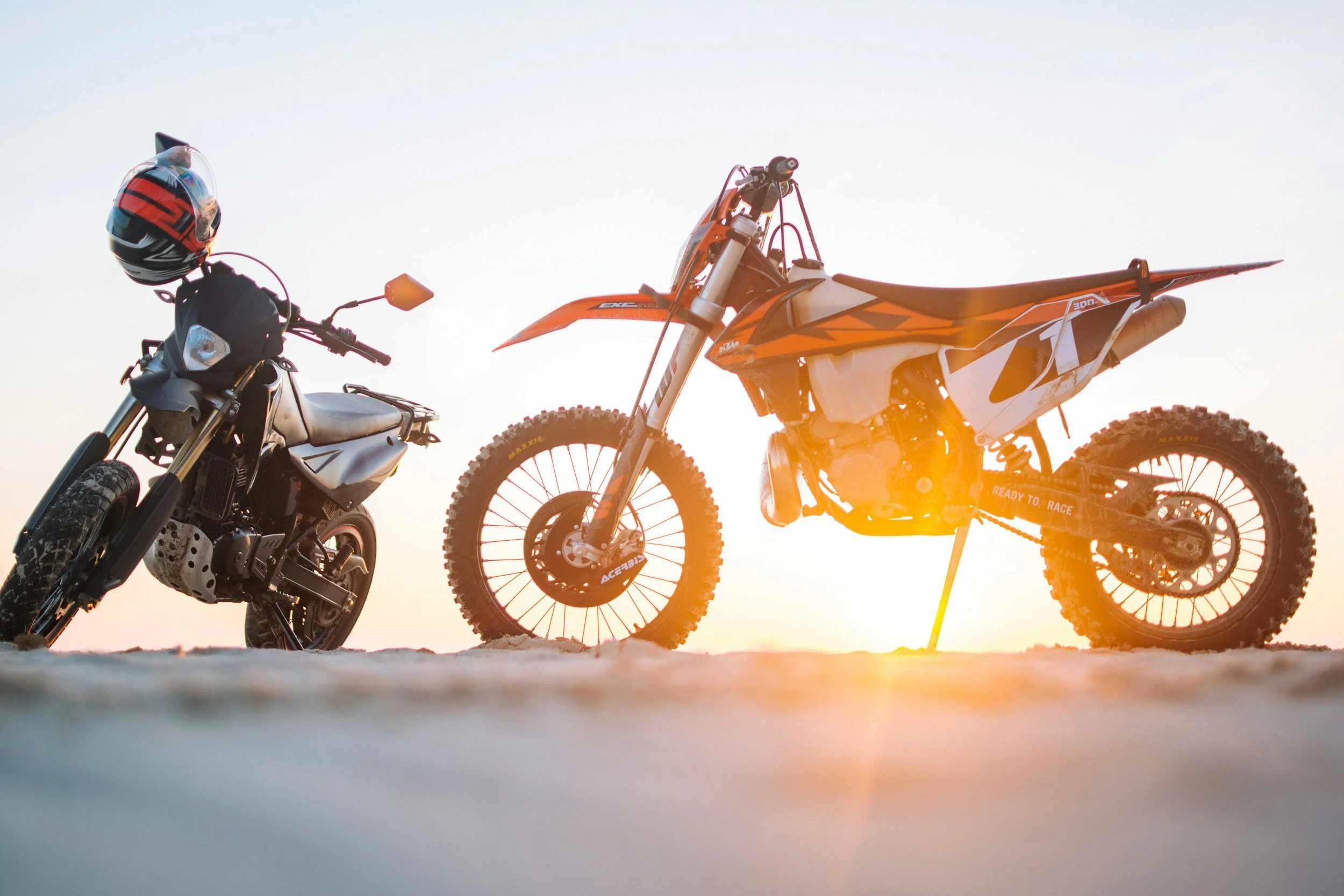 Two rides, a dirt bike and a motorcycle, are parked on a ground during sunset, with a helmet resting on the motorcycle's handlebar.