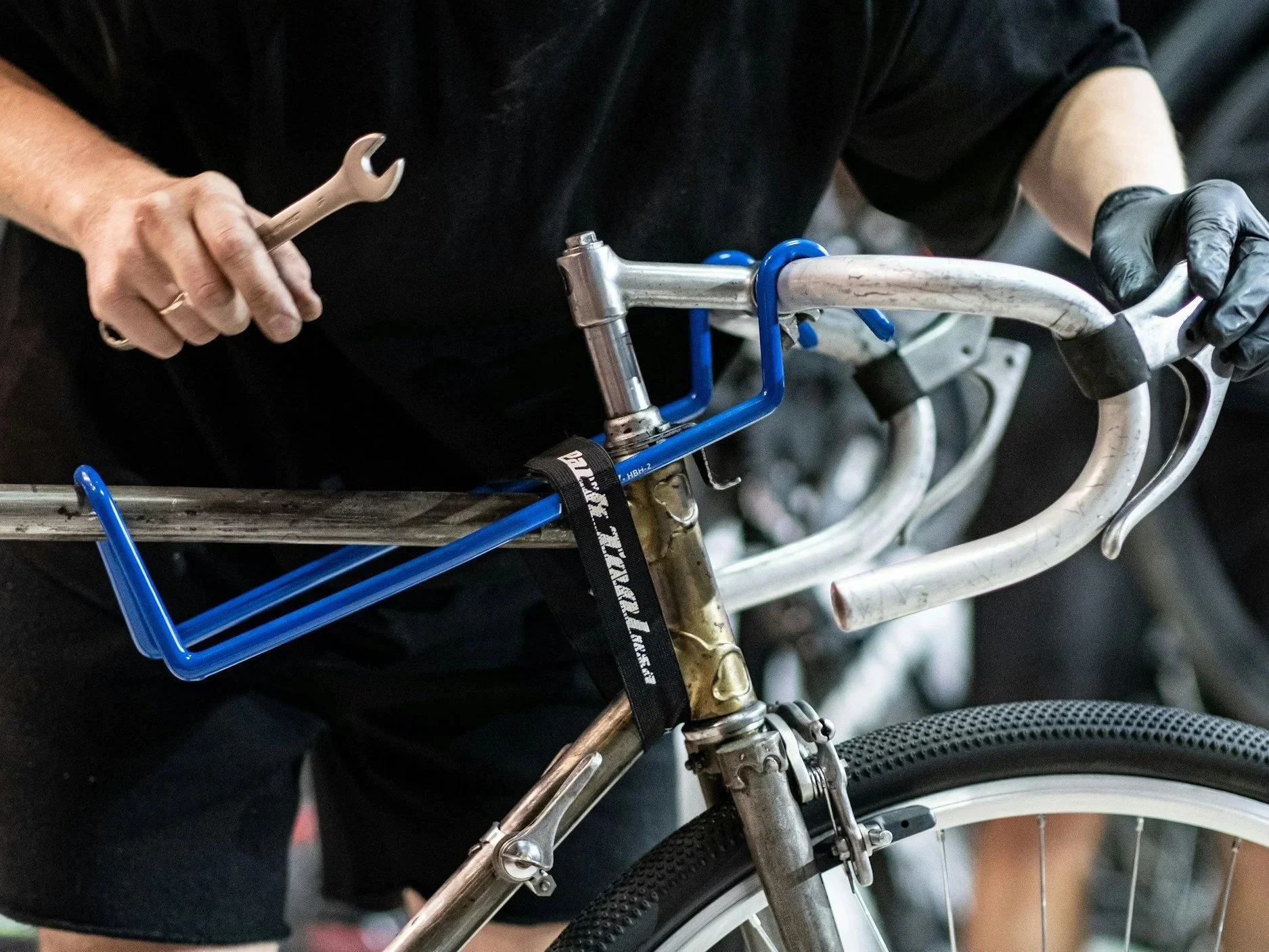 A person wearing black gloves holding the handlebar of a bicycle with a blue tool hanging from it, in a setting with other bicycles in the background.