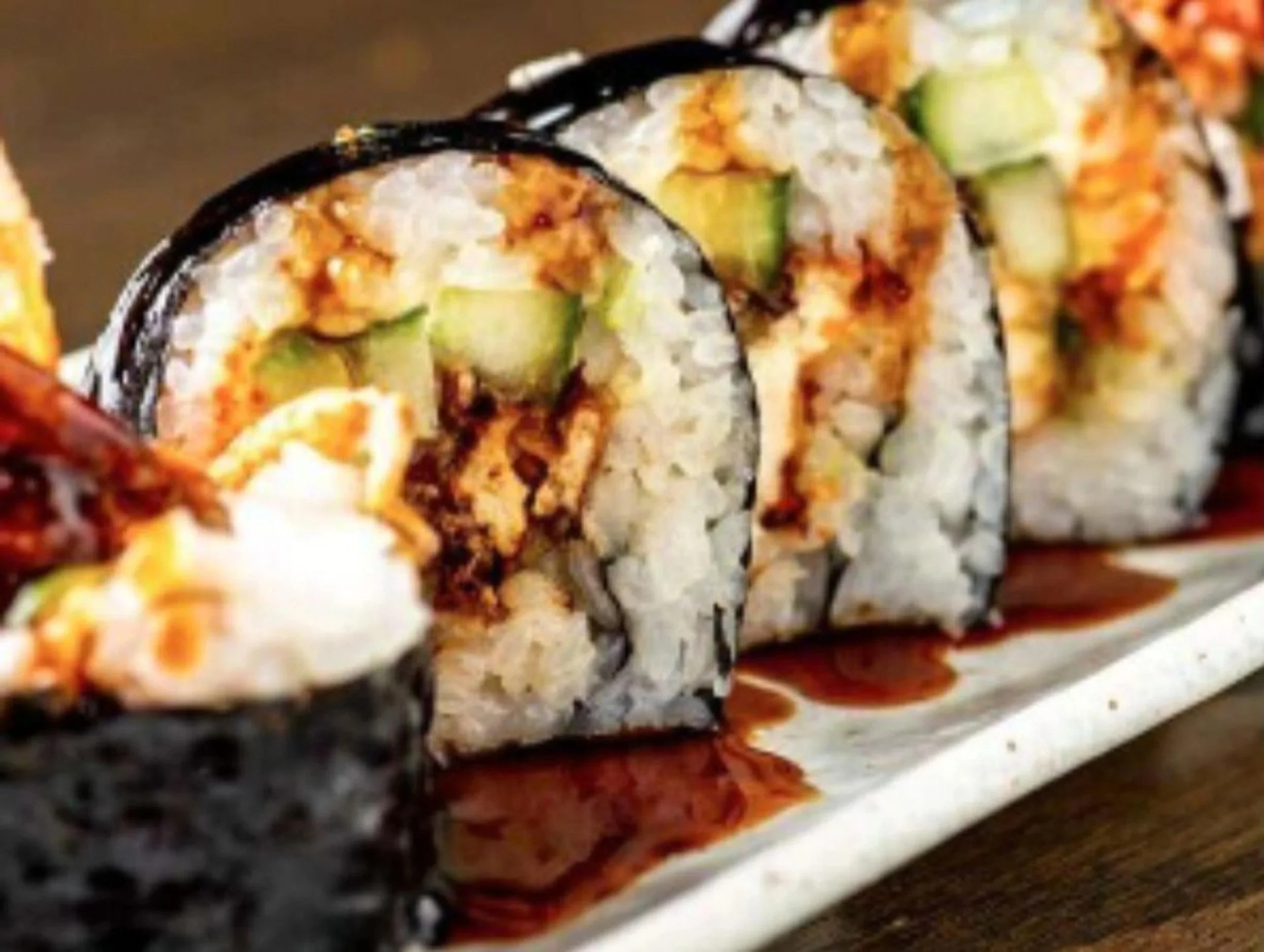 Close-up of a row of sushi rolls topped with eel, drizzled with dark eel sauce, and garnished with green avocado and cream cheese inside.