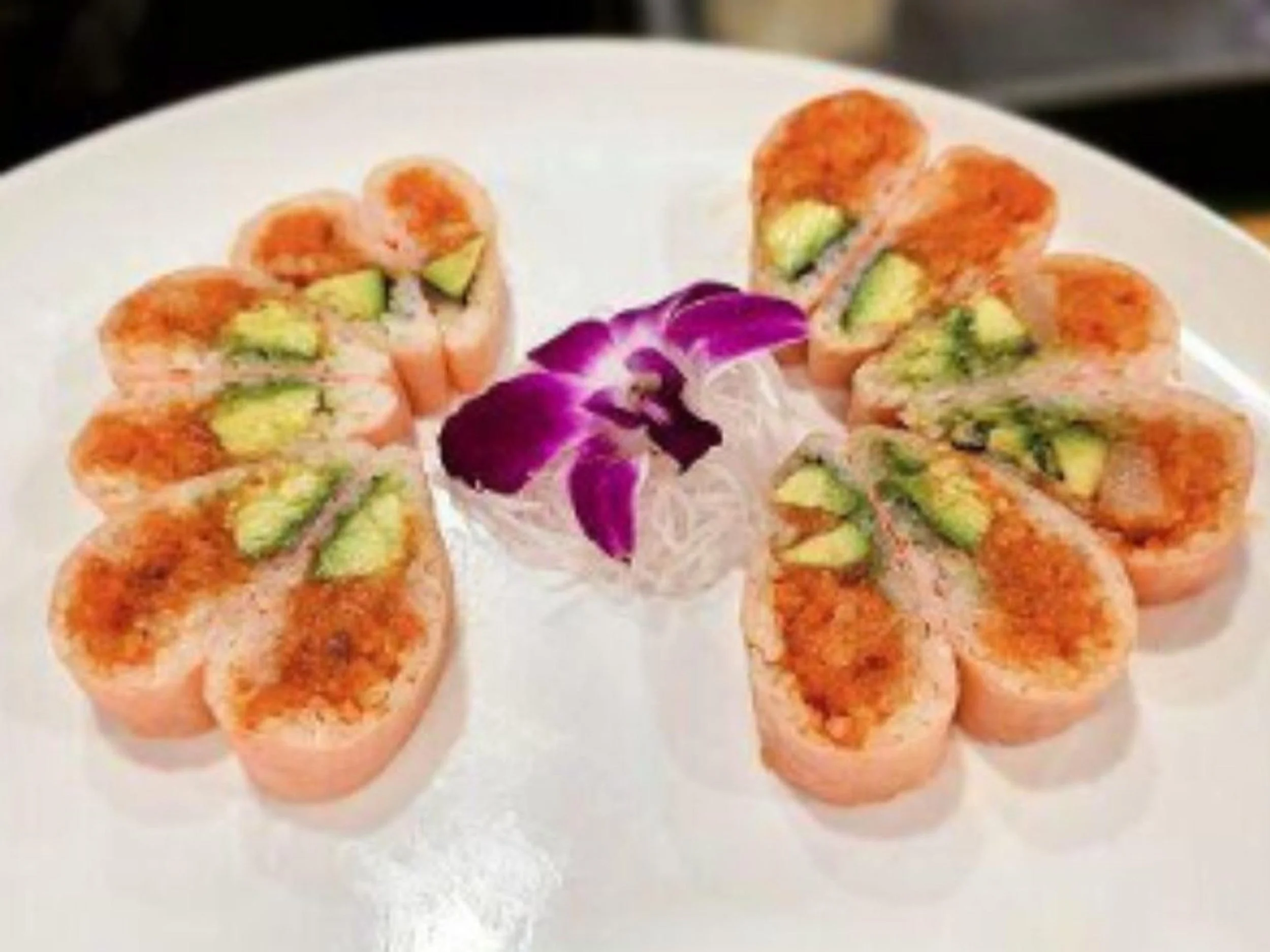 Slices of sushi arranged in a circular pattern with a purple flower in the center on a white plate.