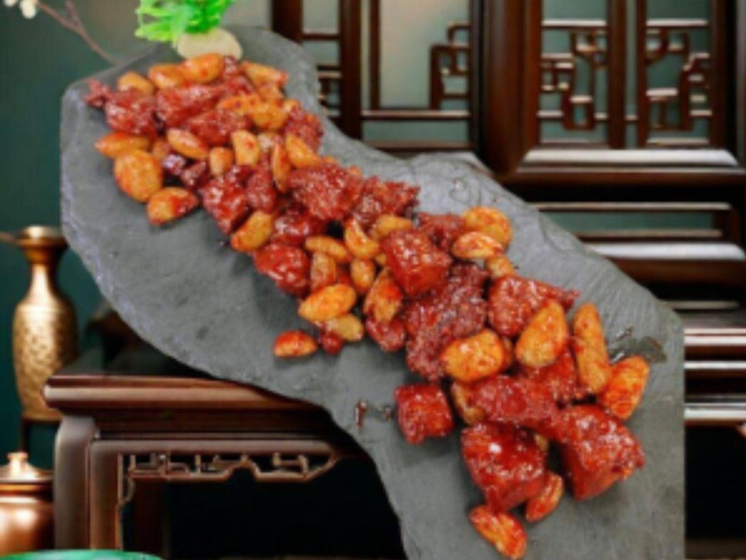 A large grey stone platter of deep-fried potato chunks coated in a red spicy sauce, placed on a table with wooden chairs in the background.