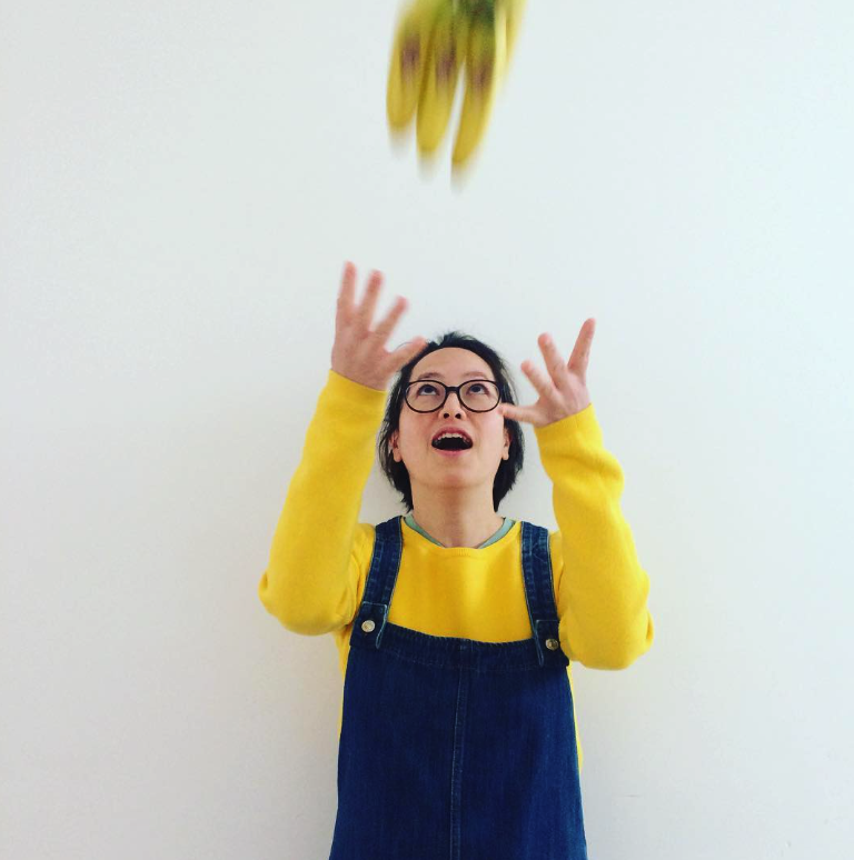 Person wearing a yellow sweater and denim overalls, throwing yellow bananas in the air with an engaging expression.