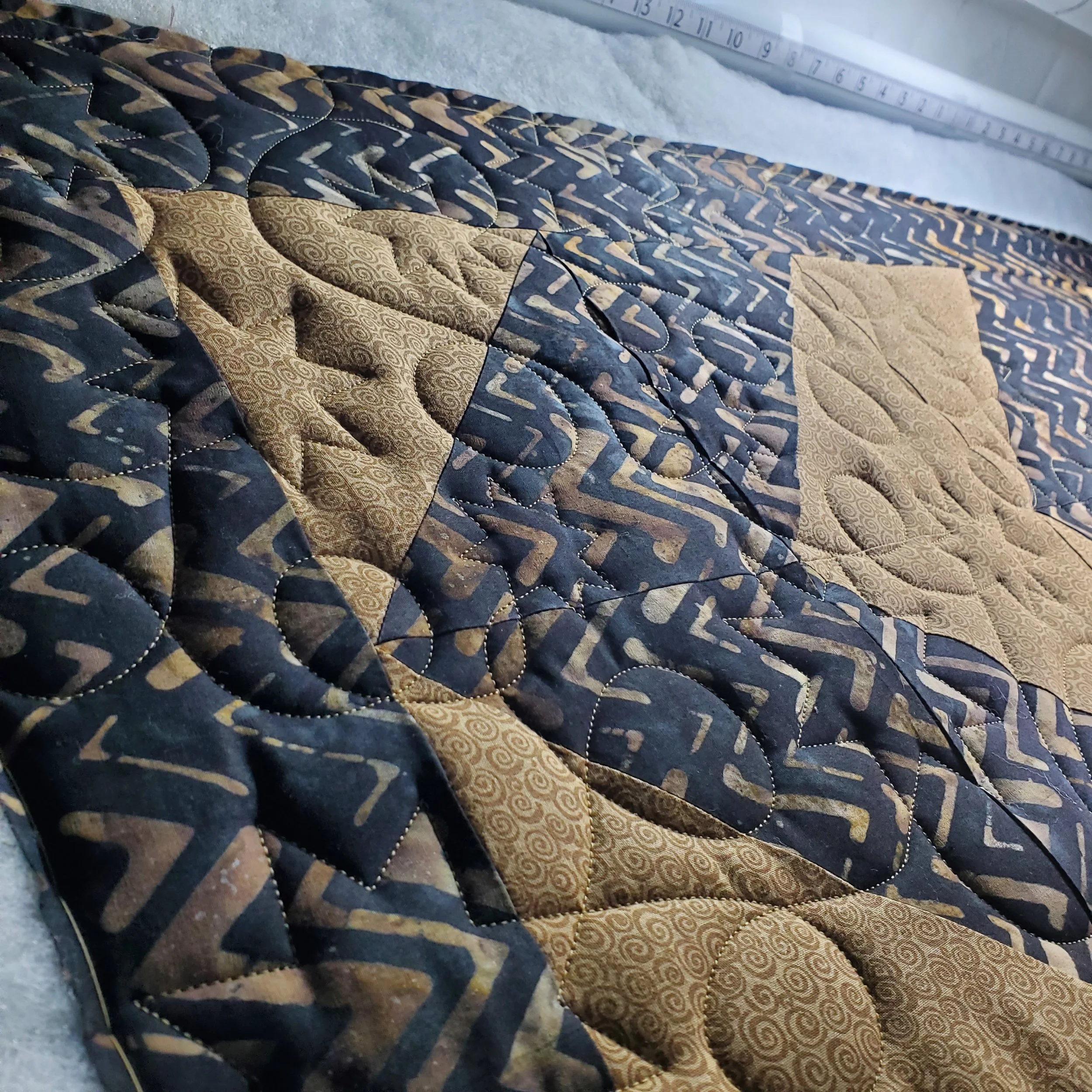 Close-up of a quilt with a patterned design in black, gold, and dark gray, placed on a surface with a white measuring tape or ruler in the background.