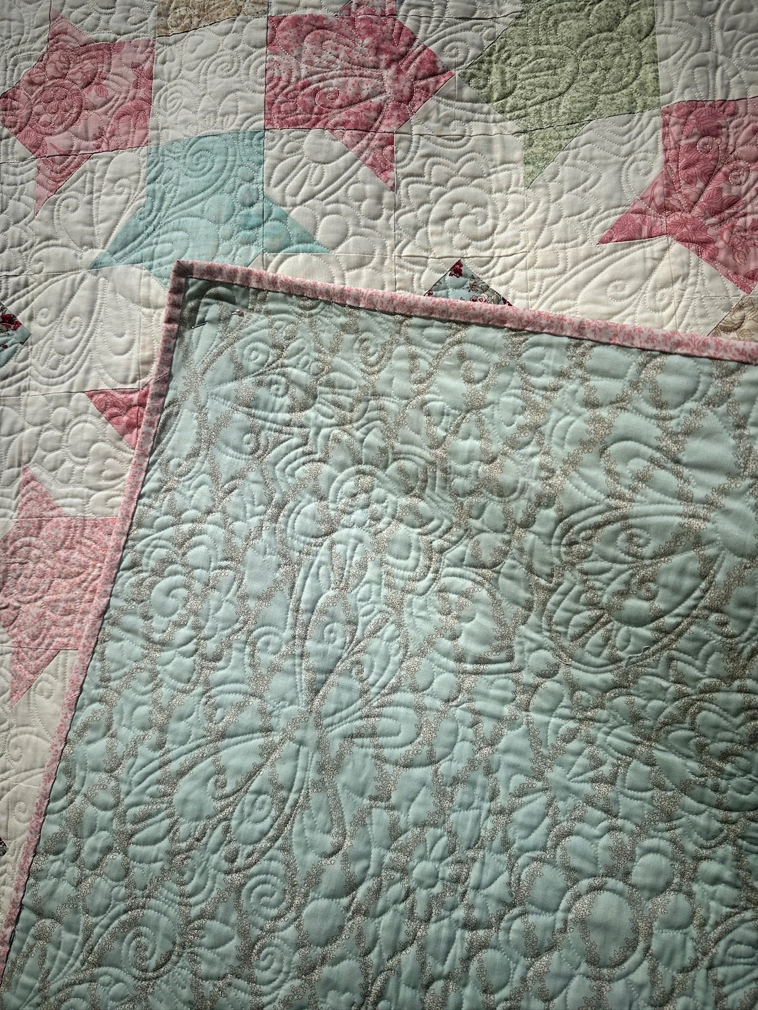 Close-up of a colorful quilt with intricate stitching, featuring pastel-colored fabric pieces with floral and swirl patterns, laid flat.