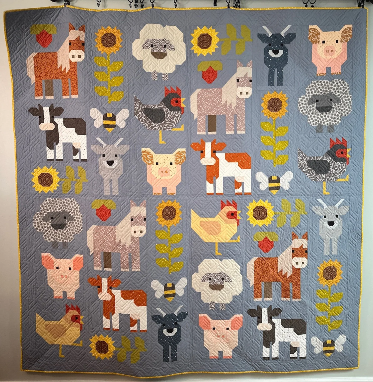 A quilt with a farm animal and sunflower motif on a blue background. The quilt features pixelated images of animals such as a horse, sheep, pig, cow, chicken, duck, rooster, and llama, along with sunflowers, bees, and strawberries, all in a colorful,