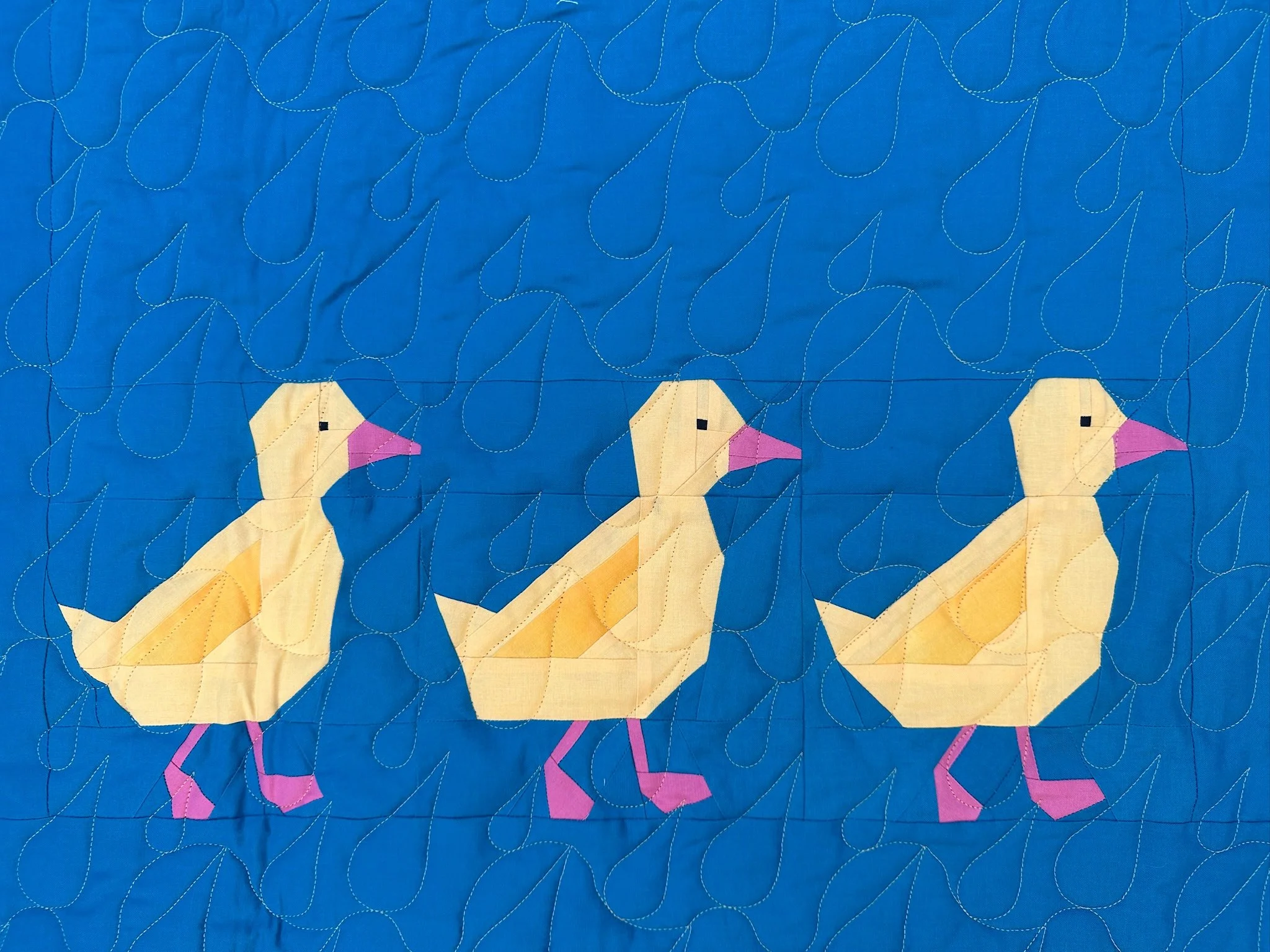 Close-up view of a colorful quilt with a blue background, featuring stylized yellow, pink, and purple birds and swirling white thread patterns.