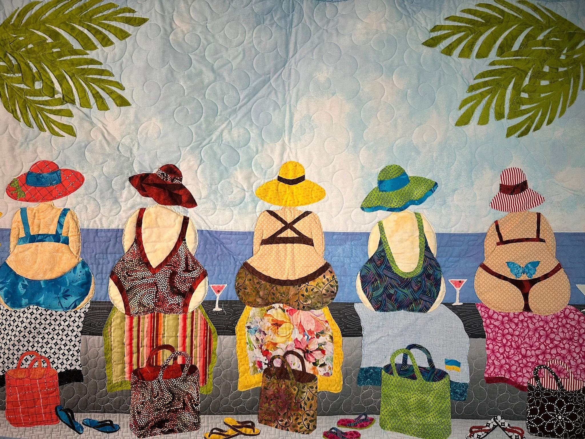 A quilted textile artwork depicting women wearing colorful hats and dresses, viewed from behind, with large green palm leaves in the background.