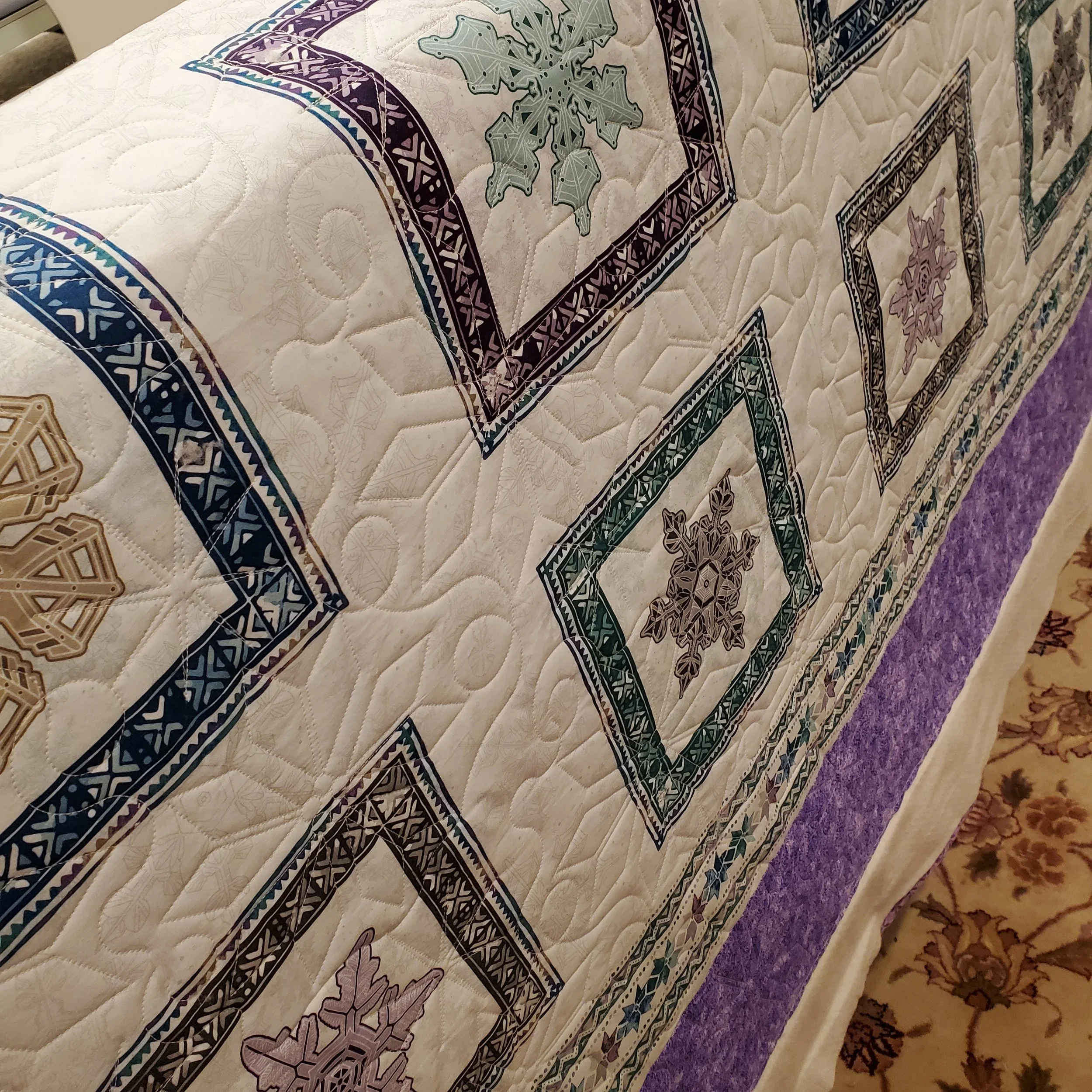 A quilt with a Christmas theme featuring snowflakes and holly leaves in various colors, decorated with intricate border patterns, lying on a surface with part of a patterned carpet visible in the corner.