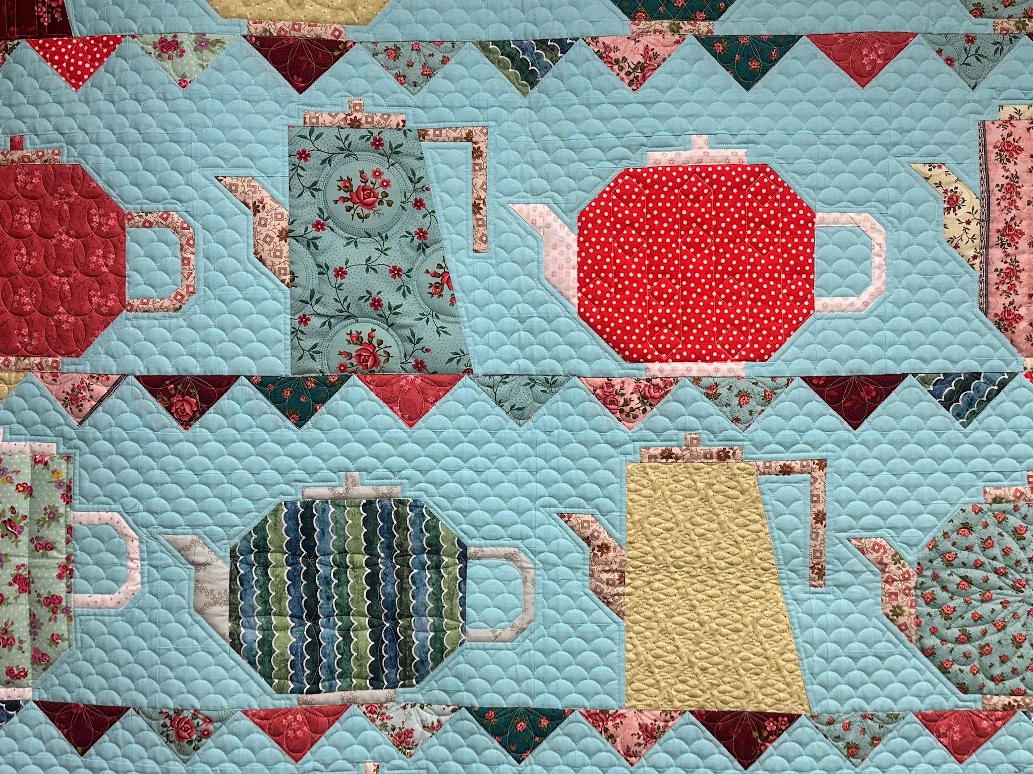 A colorful quilt with a pattern of teapots, teapots, and teapots, made from various floral and patterned fabrics, on a light blue background.