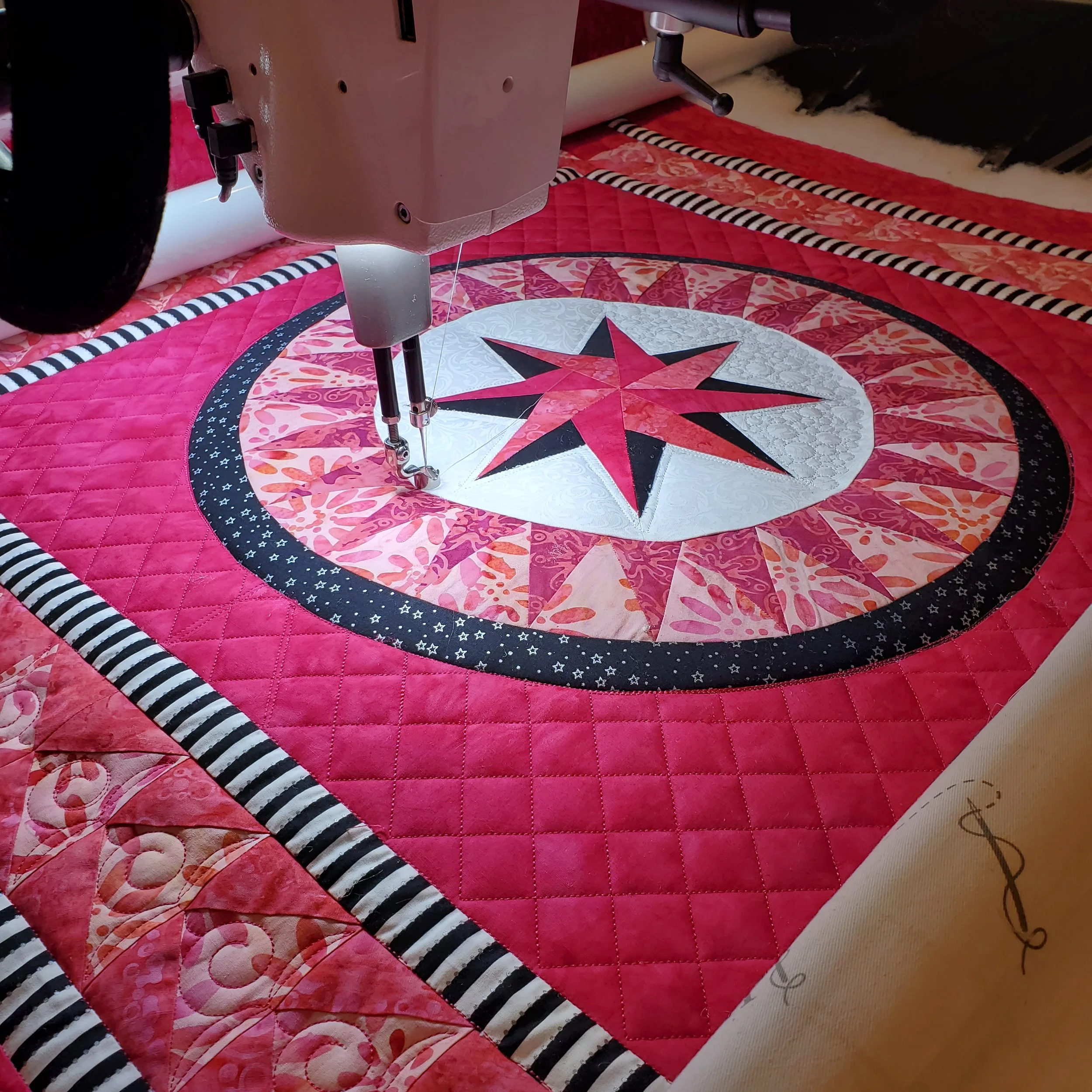 Close-up of a sewing machine quilting a pink, red, and black quilt with a star pattern, featuring striped black and white binding.
