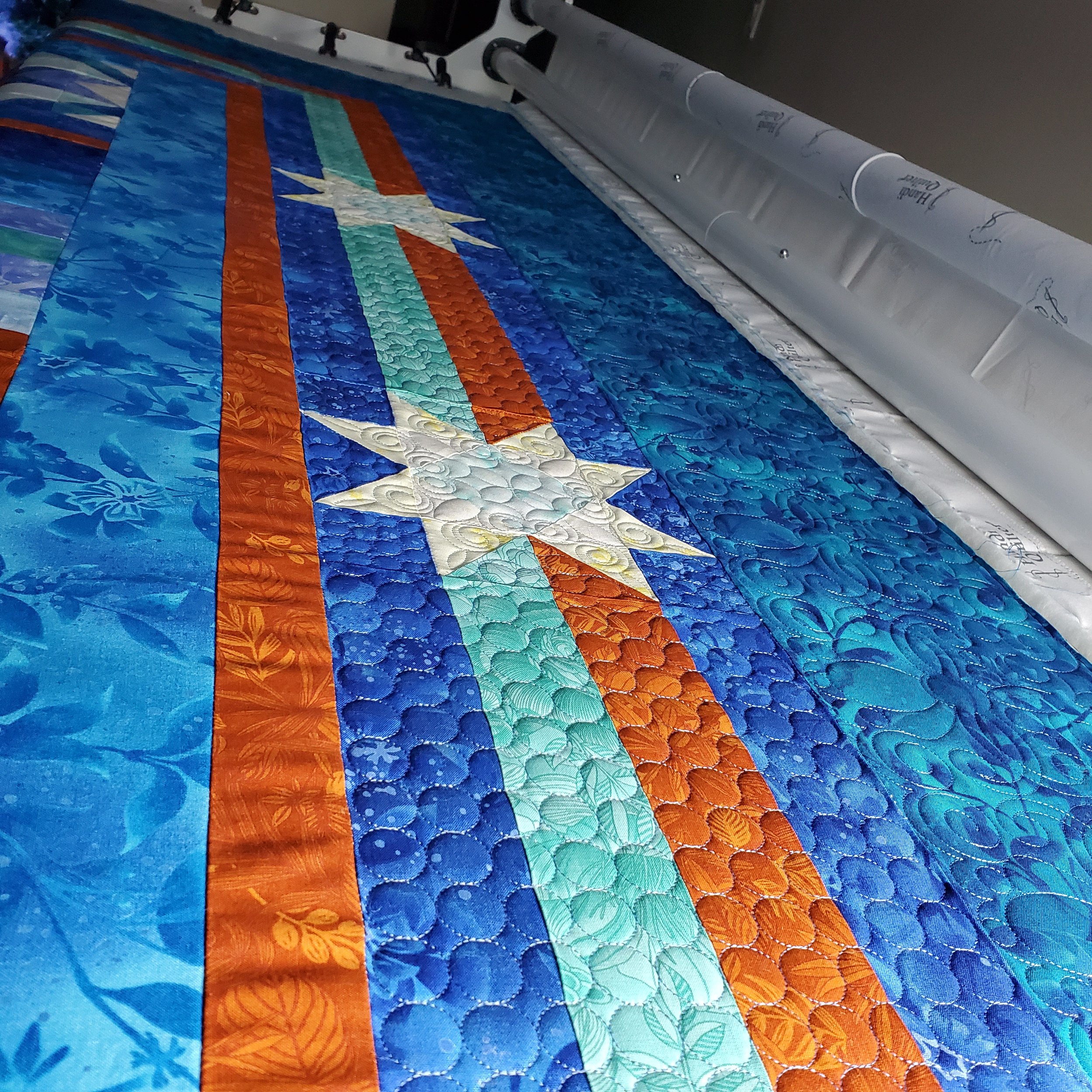 Close-up of a colorful quilt with various geometric patterns and a rainbow motif, lying under a quilting machine.