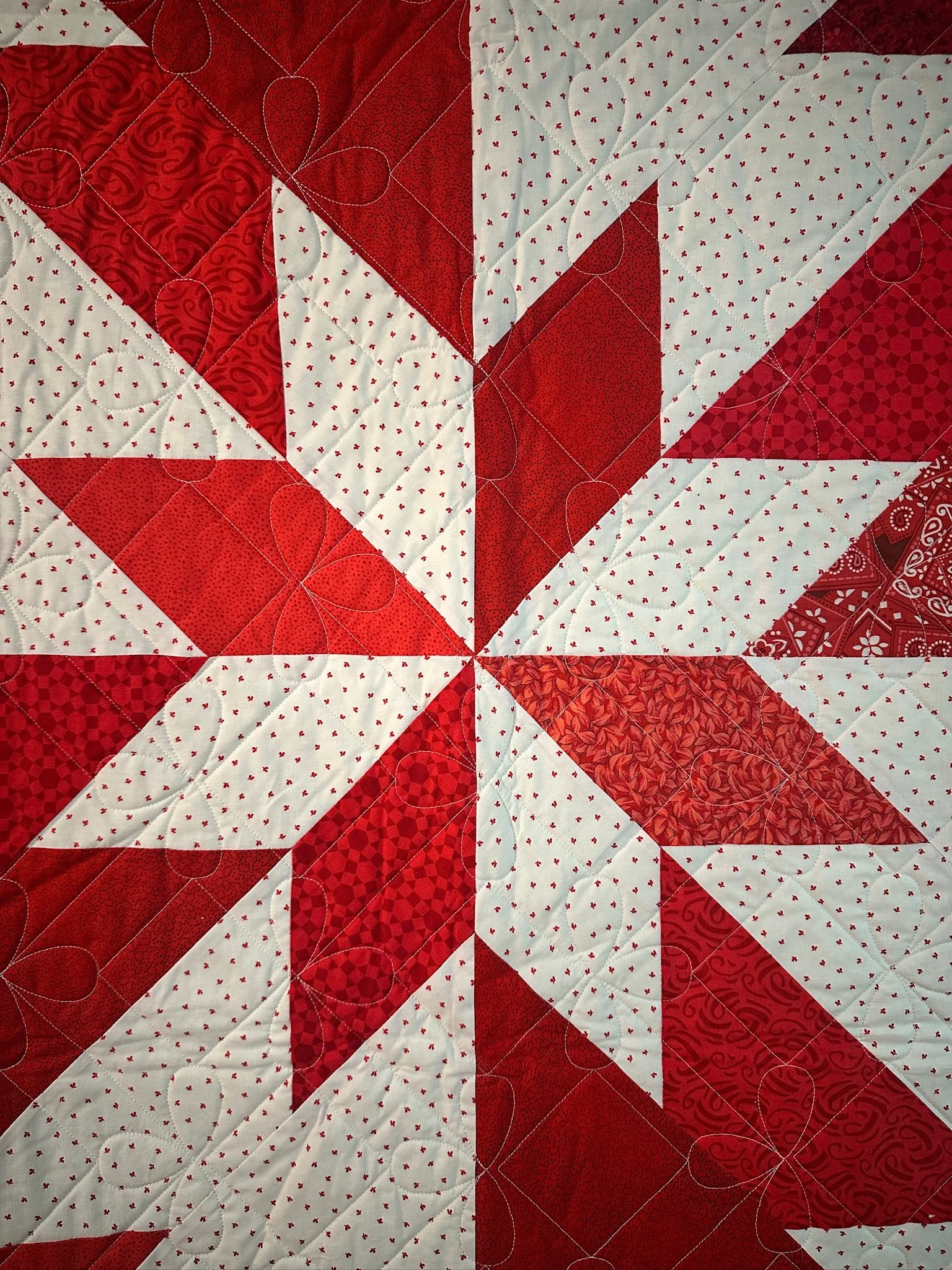 Close-up of a quilt with red and white geometric and floral patterns and white stitching details.