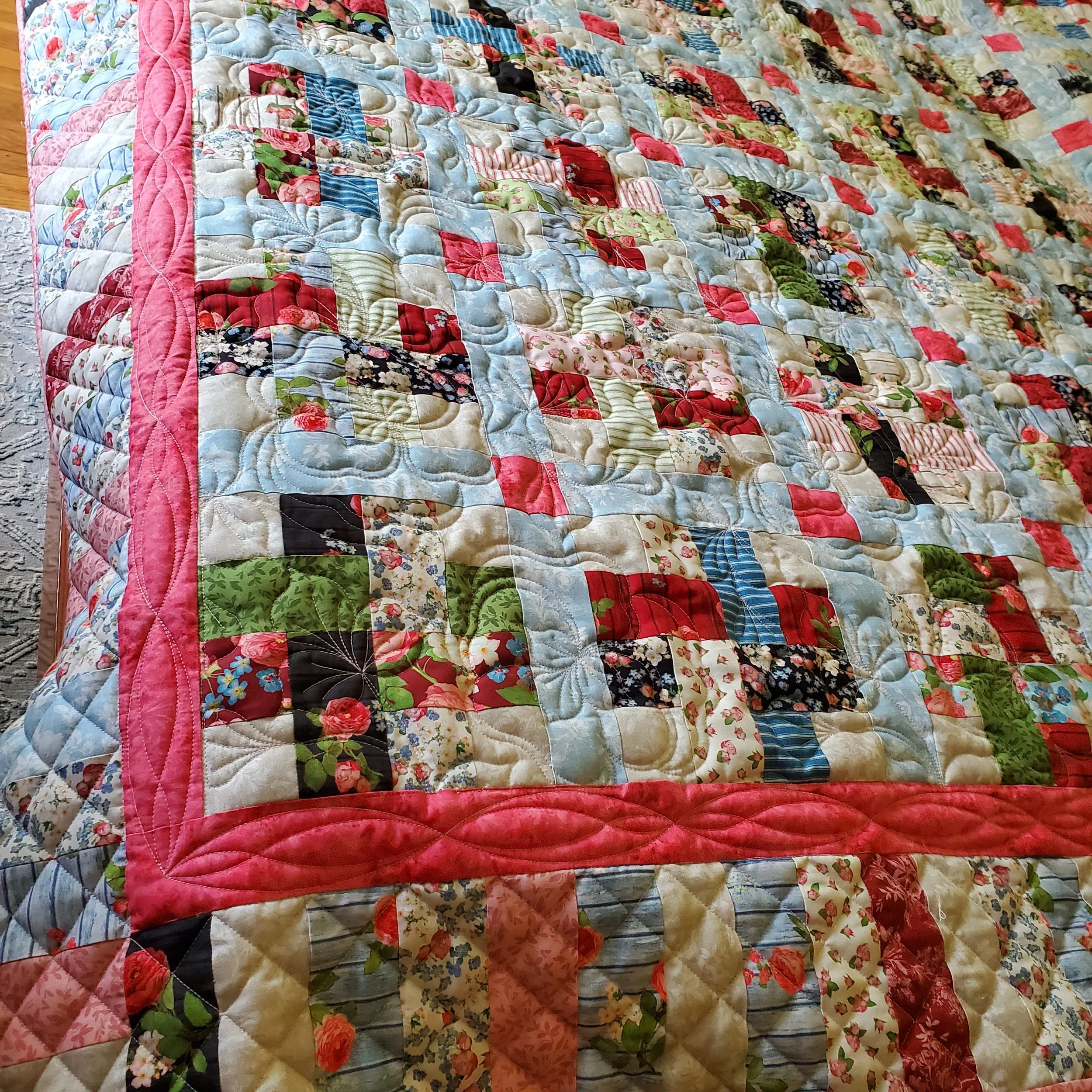 Close-up of a colorful patchwork quilt with floral and striped patterns, quilted with intricate stitching, laid on a bed with a hardwood floor nearby.