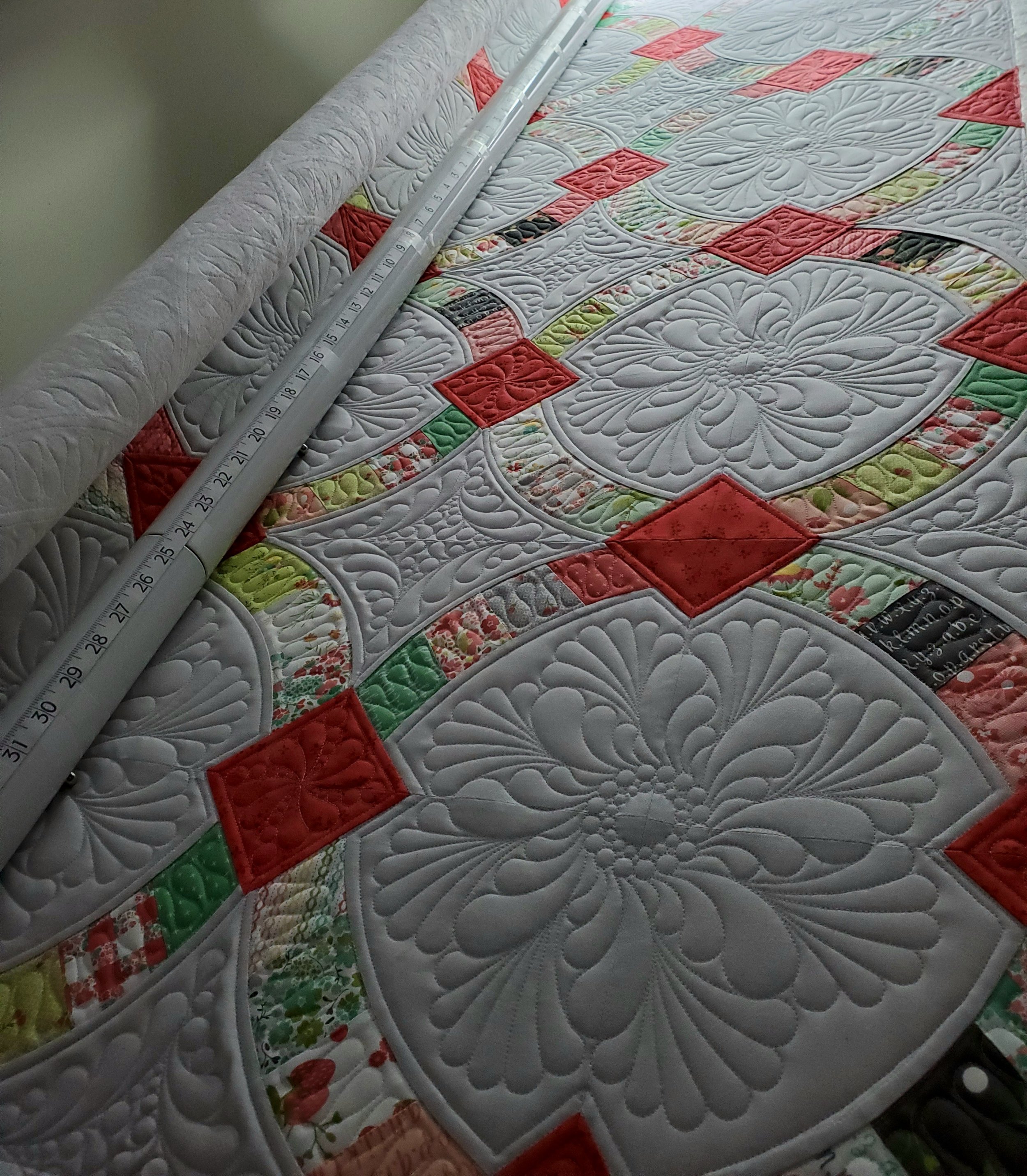 Close-up of a colorful, ornate quilt with intricate quilting patterns, partially rolled back with a ruler placed on top to show measurement.