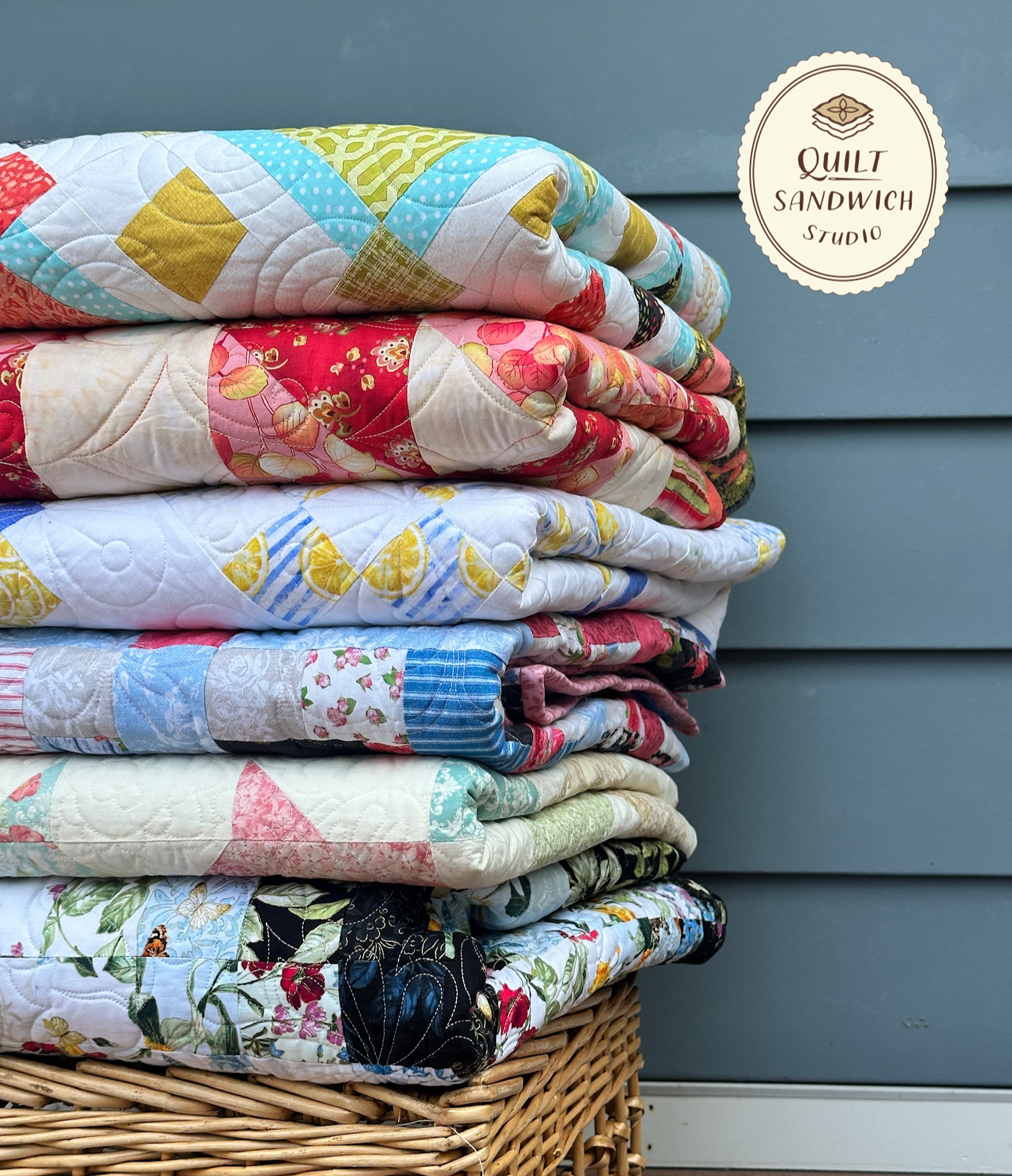 A stack of colorful, longarm quilted quilts on a wicker basket . There is a Quilt Sandwich Studio logo in the top right corner.