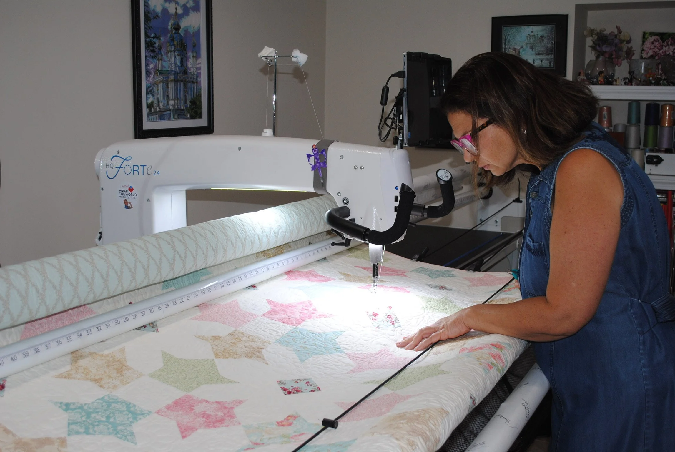 A woman  sewing a quilt on a computerized longarm quilting machine in a room with framed pictures and a shelf with vases and flowers.