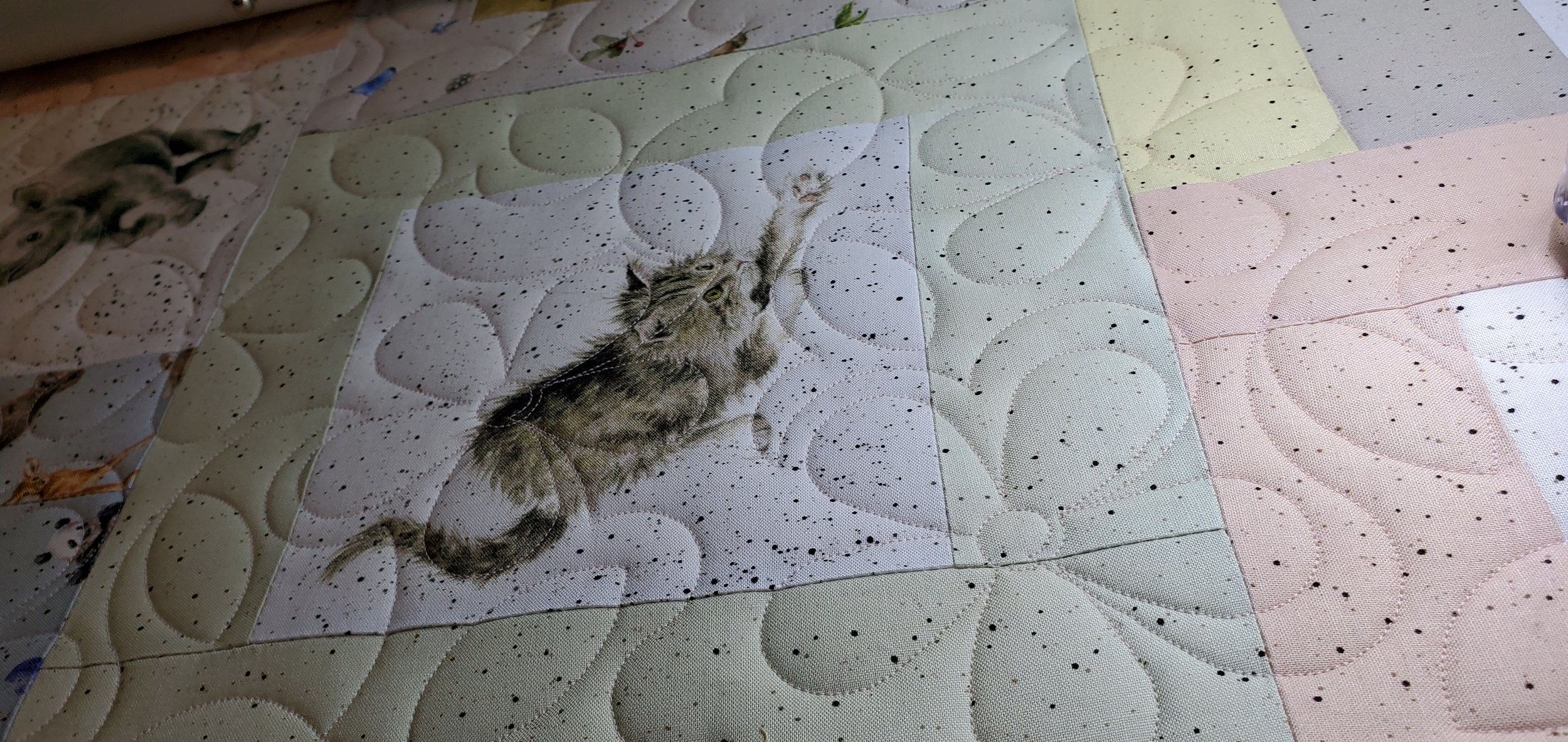 A quilt with a cat design, showing a gray and brown tabby cat stretching with paws outstretched, on a patchwork pattern of various colors and decorative stitching.