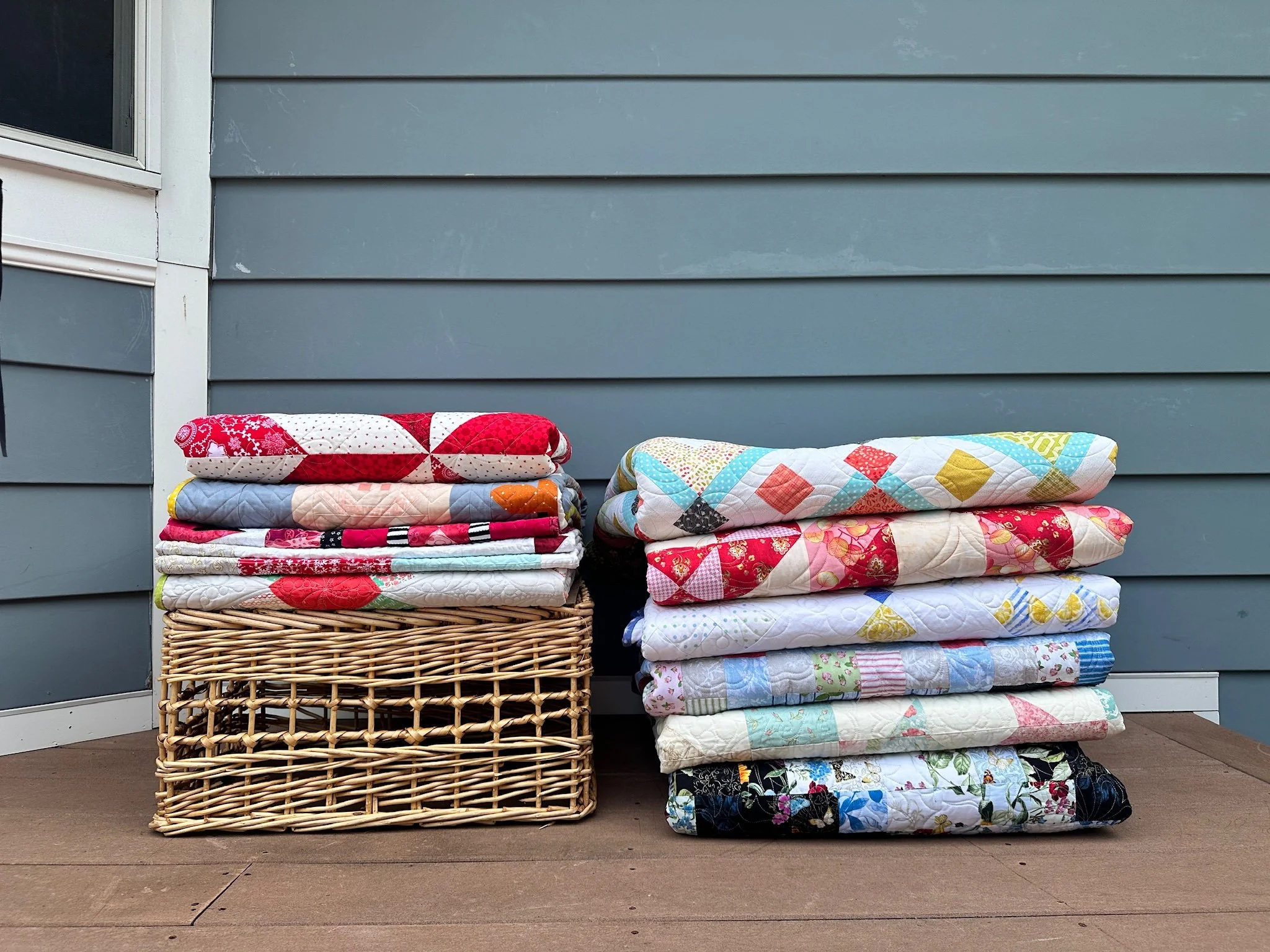 Stacks of colorful long arm quilted blankets and quilts on a wicker basket and on the ground, placed outside against a blue house wall with siding and a window in North Shore MA.  Longarm service inMarblehead MA