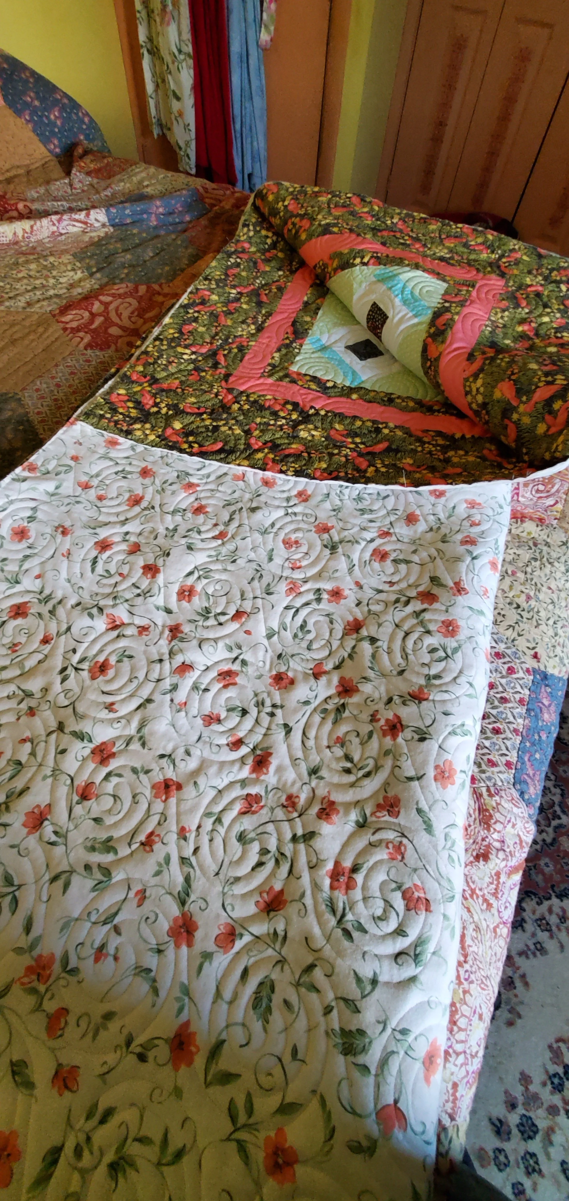 A bed with floral and patterned quilts, with a folded quilt on top, in a bedroom with colorful curtains and wooden wardrobe.